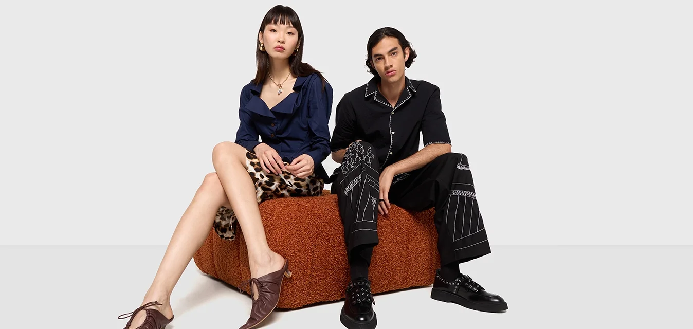 Dos personas están sentadas en un asiento texturizado marrón. Una lleva una camisa azul y pantalones cortos estampados; la otra, una camisa negra y pantalones rayados.