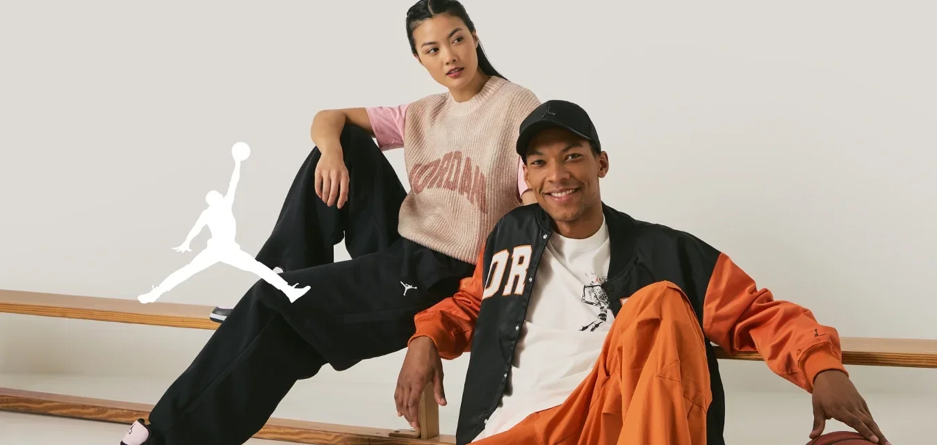 Dos individuos se sientan de manera casual en un banco de madera. El hombre lleva una chaqueta naranja y negra, una camiseta blanca, pantalones naranjas y zapatillas grises. La mujer viste un top de punto rosa y beige con pantalones negros y zapatillas negras.