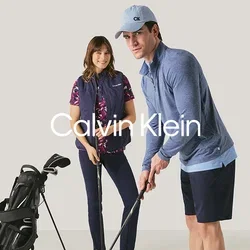 Hombre y mujer preparándose para jugar al golf en interiores con palos de golf y una bolsa de golf sobre un fondo neutro.