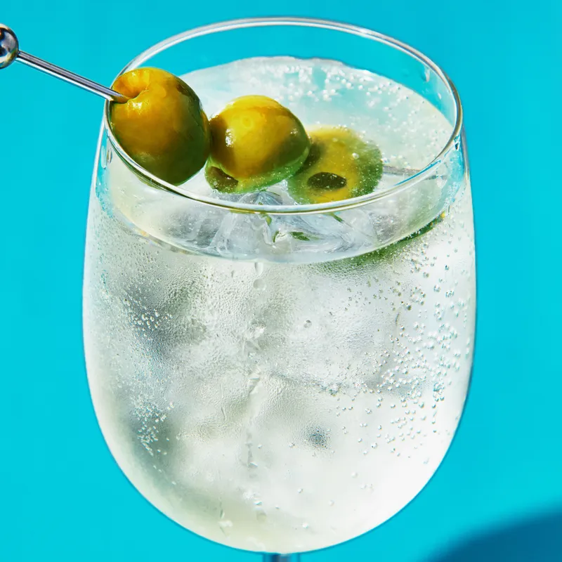 A bubbly dirty martini spritz served over ice in a stemmed wine glass and garnished with three Castelvetrano olives on a cocktail pick.