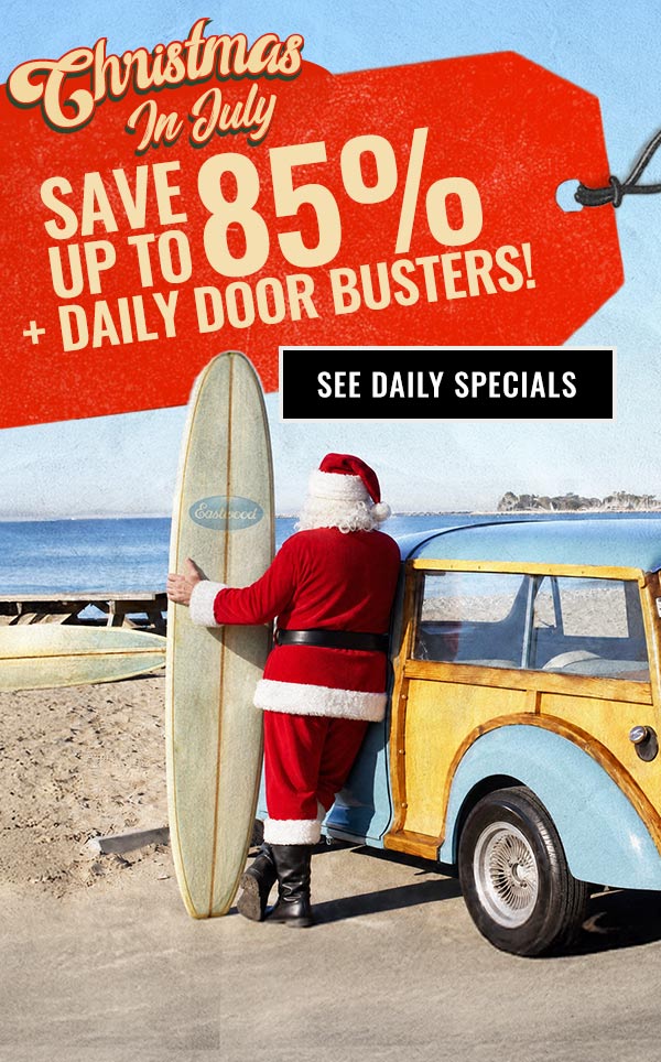 Santa stands beside a vintage woody wagon holding a surfboard on the beach with a large price tag announcing "Christmas in July – Save Up to 85% + Daily Door Busters."