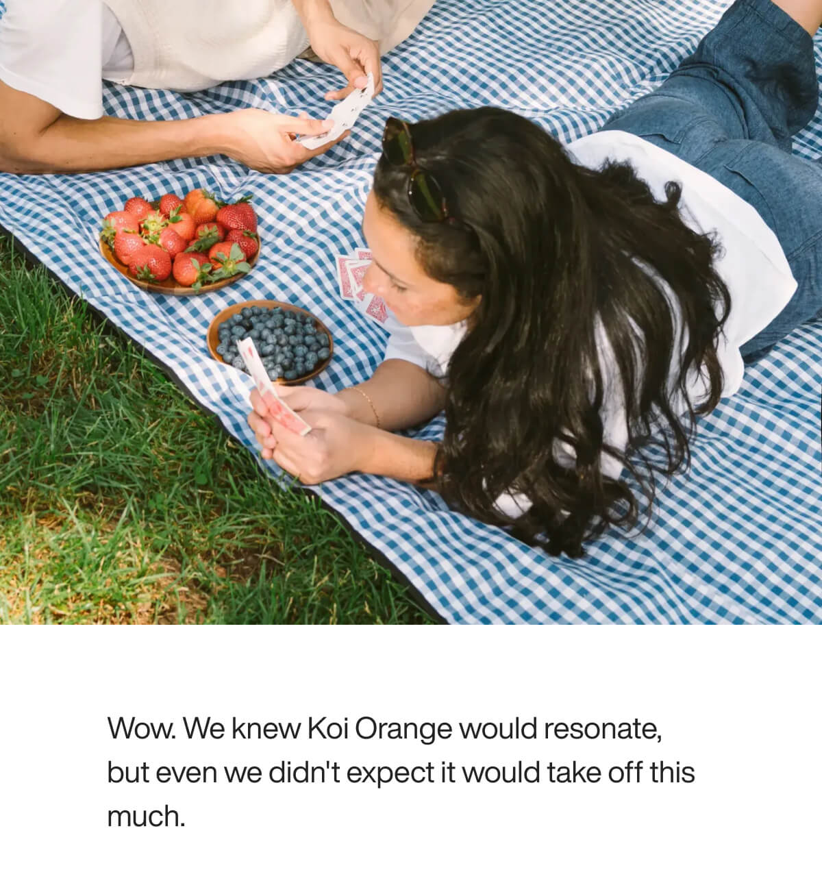 "Two people play cards on a picnic blanket with strawberries and blueberries, enjoying a sunny day outdoors."