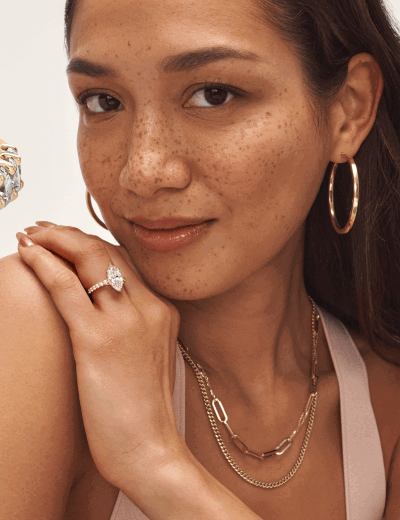 Woman wearing gold hoop earrings, layered gold necklaces and a marquise diamond ring resting her hand on her shoulder.