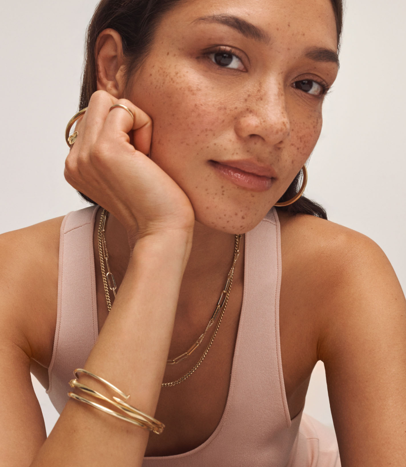 Woman wearing layered yellow gold necklaces, hoops, bangles and rings in a soft blush tank top.