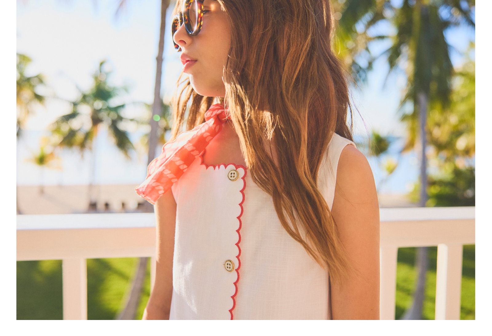 Girl in white scallop-trim dress with coral neck scarf and sunglasses, standing on a balcony with palm trees in the background