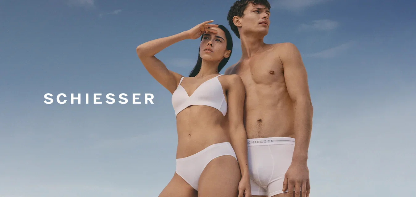 Jonge vrouw in witte beha en ondergoed die haar ogen beschermt tegen de zon naast een bovenlichaam naakte man in witte boxershorts tegen een blauwe lucht achtergrond.