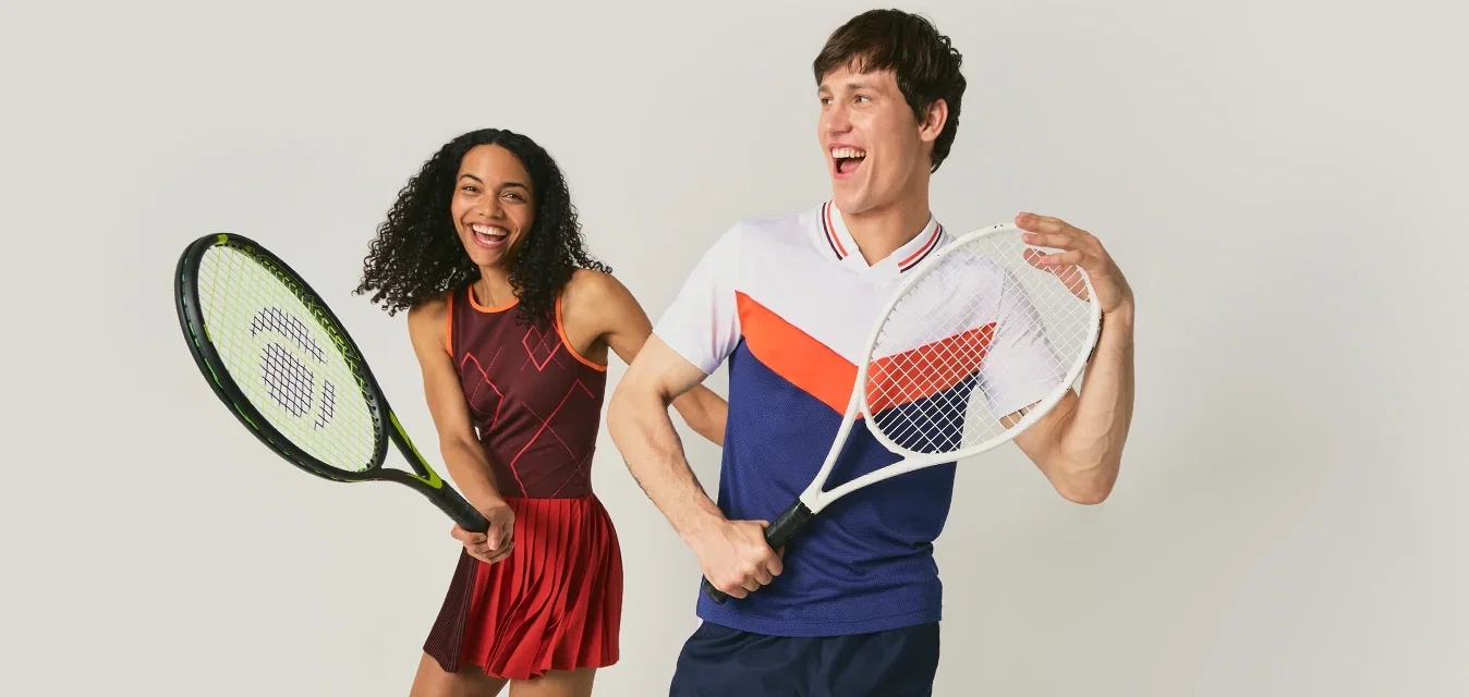 Un hombre y una mujer sonrientes con ropa de tenis posan con raquetas, de pie frente a un fondo claro y sencillo.