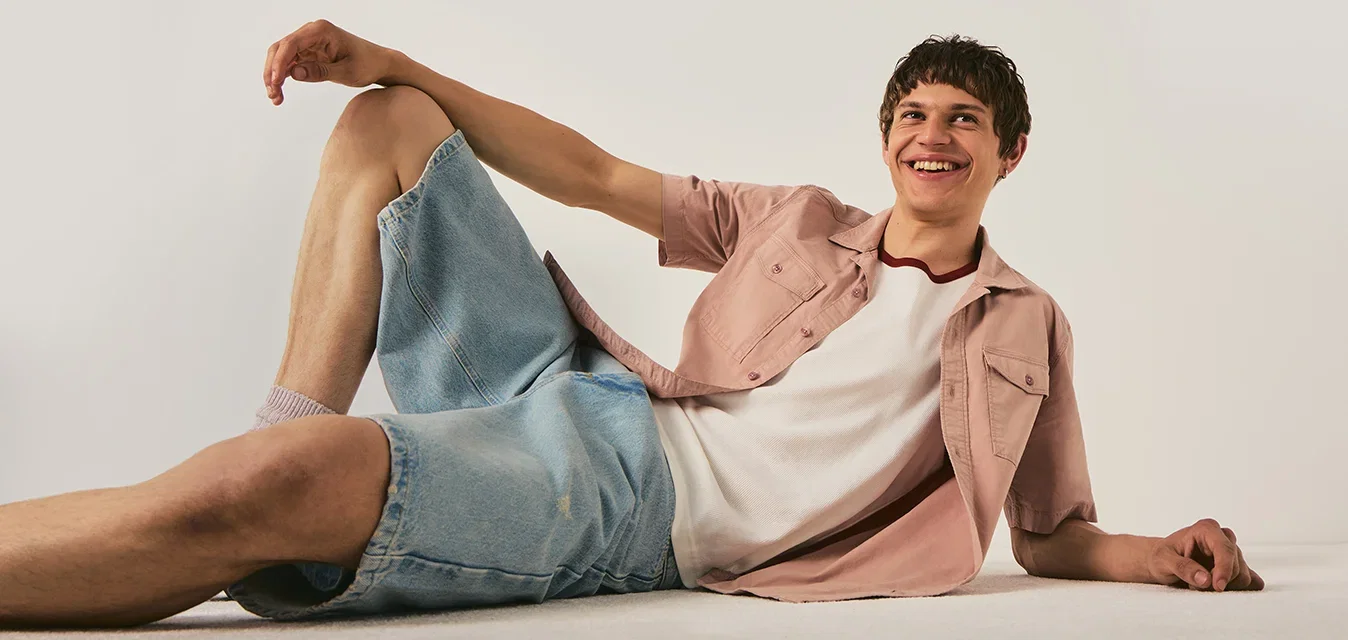 Joven con pantalones cortos vaqueros y camisa rosa recostado en el suelo sonriendo contra un fondo blanco liso.