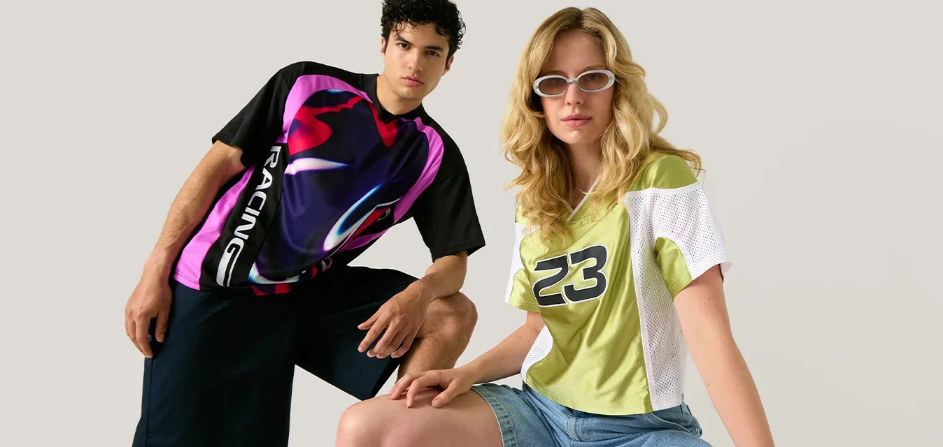 Dos adultos jóvenes llevan ropa deportiva informal; uno está sentado en un taburete con gafas de sol, el otro está de pie detrás con un fondo liso.