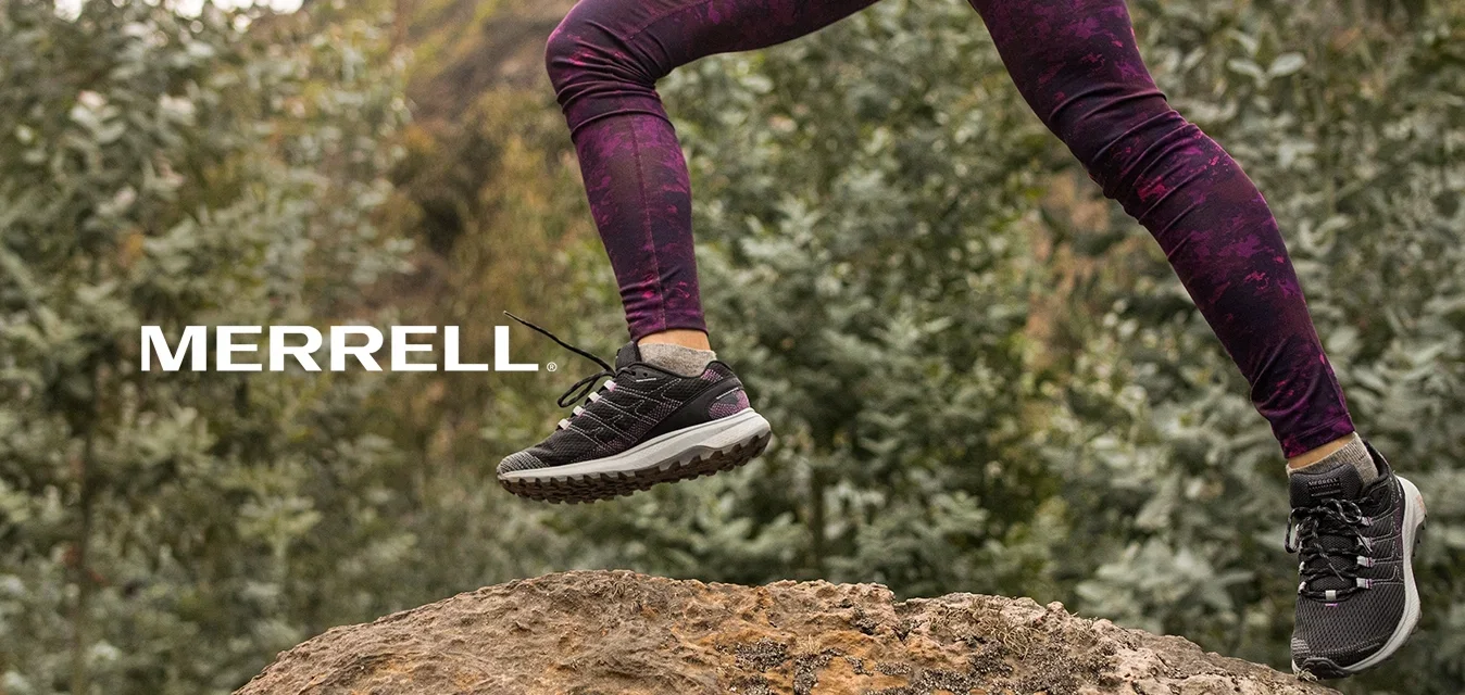 Persona vestida con leggins morados y zapatillas negras saltando sobre una gran roca en un área boscosa al aire libre.