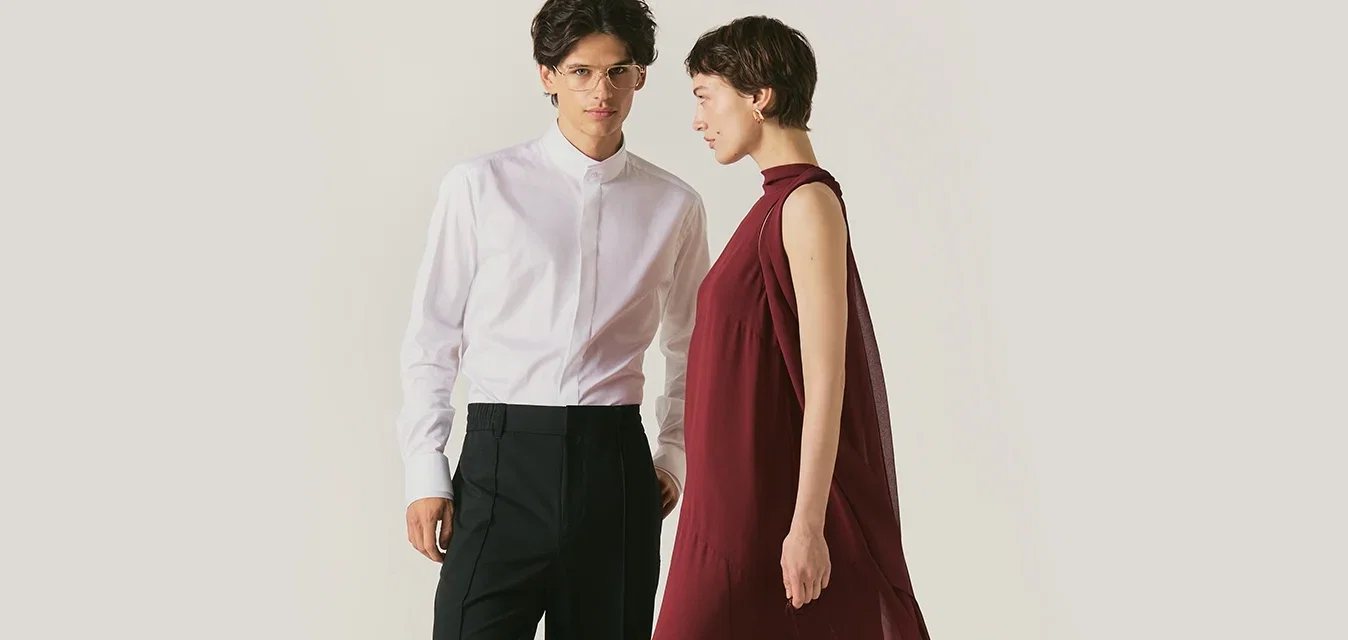 Giovane uomo in camicia bianca e pantaloni neri accanto a una donna in un abito bordeaux fluido che tiene una piccola borsa, entrambi posano.