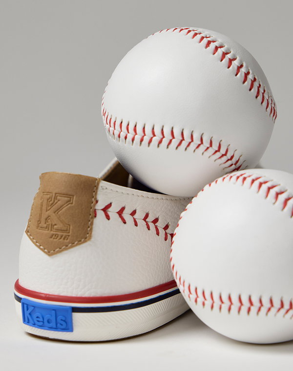 A closeup of the back of the Keds Champion Pennant Leather Lace-Up in off white highlighting the sneaker’s embossed leather home plate heel tab, red baseball stitching, and blue Keds logo. There are two baseballs sitting with the sneaker, one to its right and the other nestled in its top.