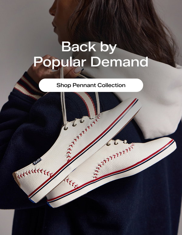 A view of the back of a woman wearing a hooded varsity jacket in navy, white, and red. She is holding a pair of Keds Champion Pennant Leather Lace-Ups in off white over her shoulder by the laces. The image highlights the sneakers’ rich, textured leather and red baseball stitching. Text over the image says Back by Popular Demand. Shop Pennant Collection.