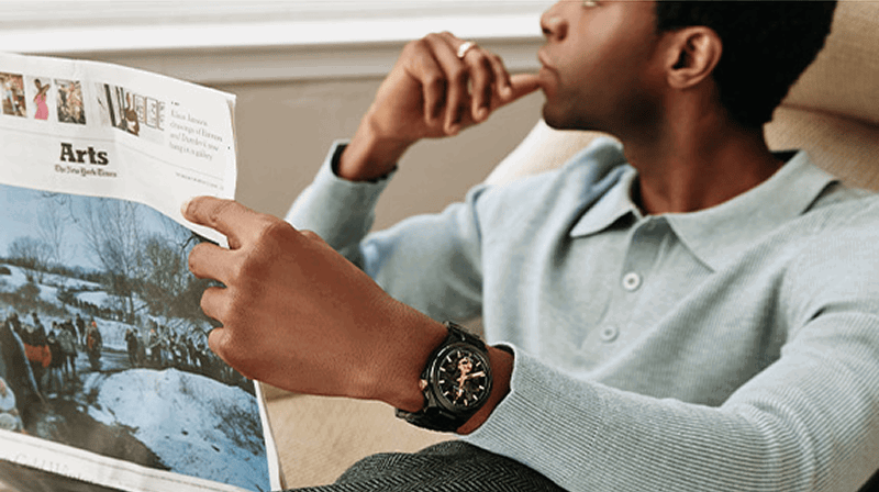 Man reading a newspaper while wearing a black Bulova Maquina automatic watch with gold-tone details.