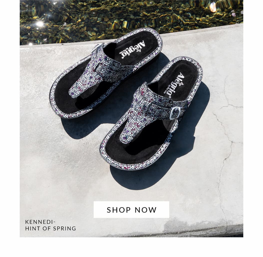 Pair of Alegria Kennedi Hint of Spring mosaic-patterned thong sandals with side buckles resting on a stone surface with a Shop Now button.