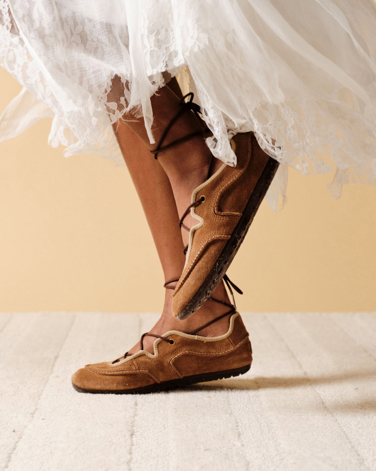 Person's lower legs in white lace skirt, wearing the Quill Ballet Trainer on a wooden floor.