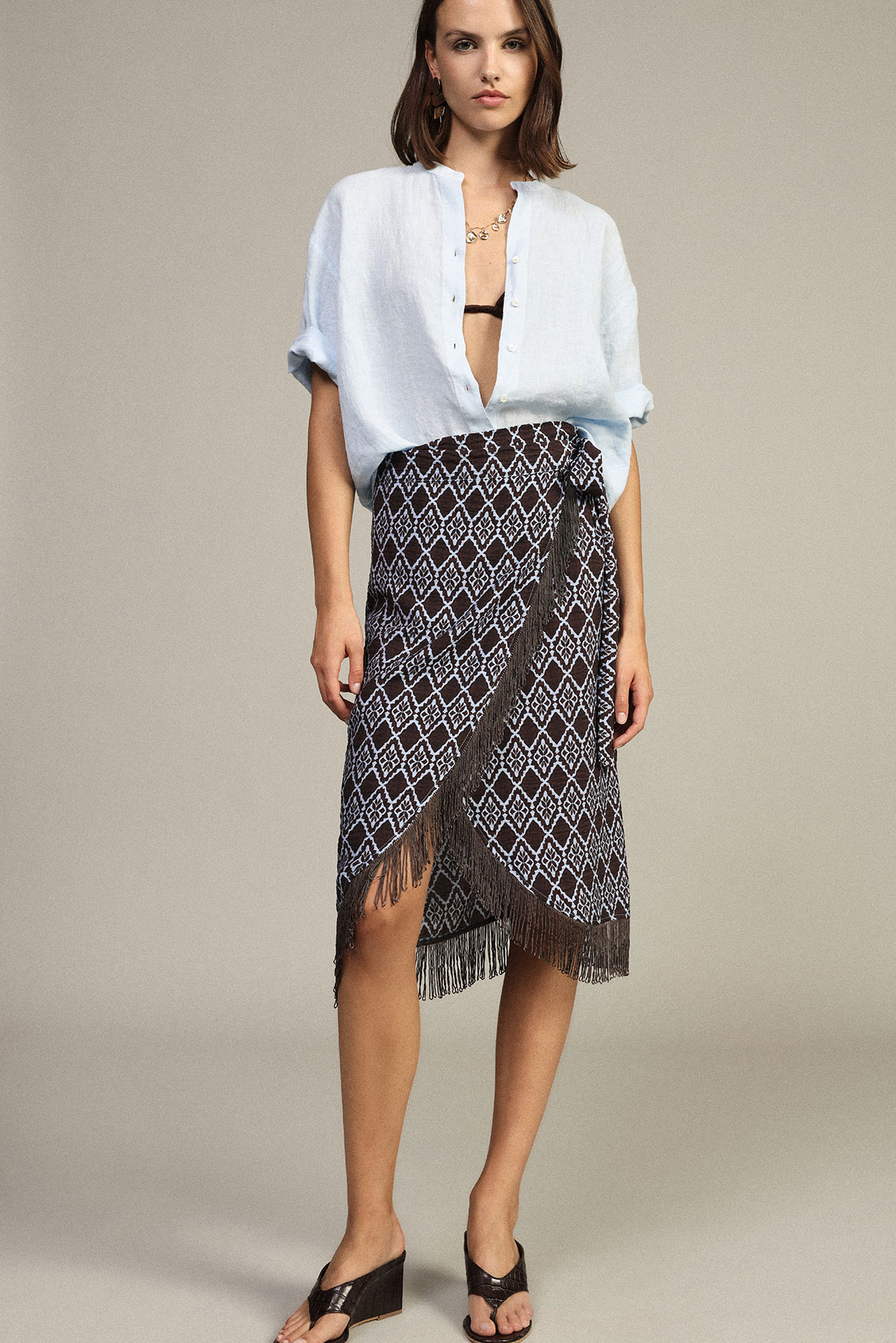 Woman modelling light blue open shirt with black bikini top and patterned wrap skirt with fringe, black wedge sandals
