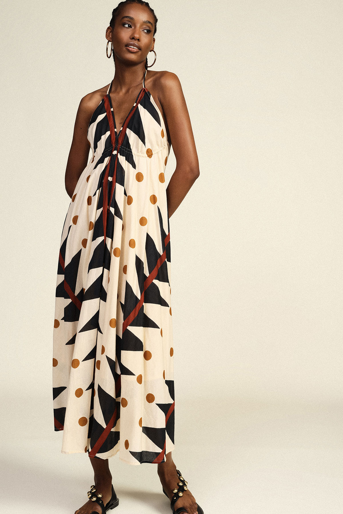Model modelling sleeveless halter maxi dress with black geometric and mustard polka-dot pattern, hoop earrings and sandals