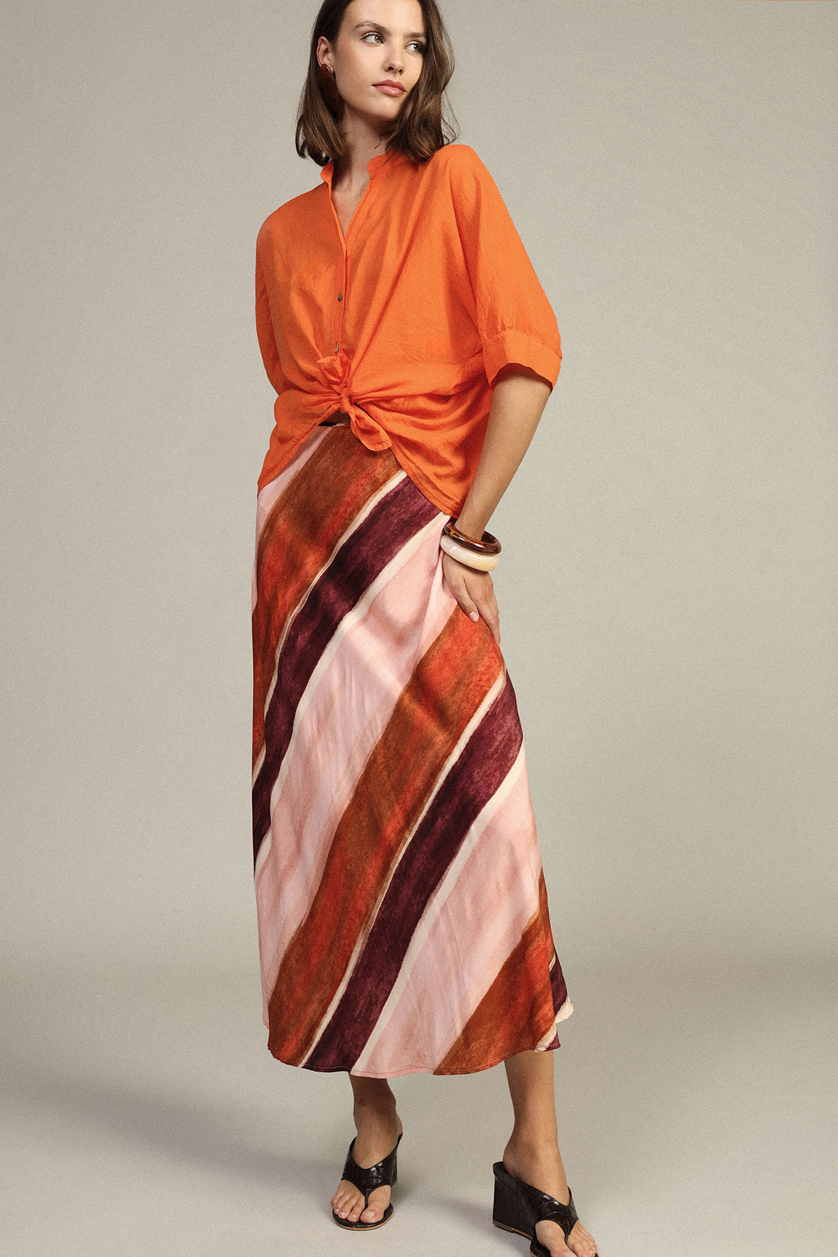Woman modelling tied orange blouse and diagonal striped maxi skirt with black sandals on neutral background