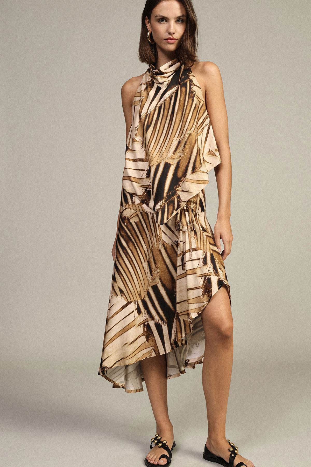 Woman wearing sleeveless halter-neck midi dress with brown tropical leaf print and asymmetric hem, black studded sandals