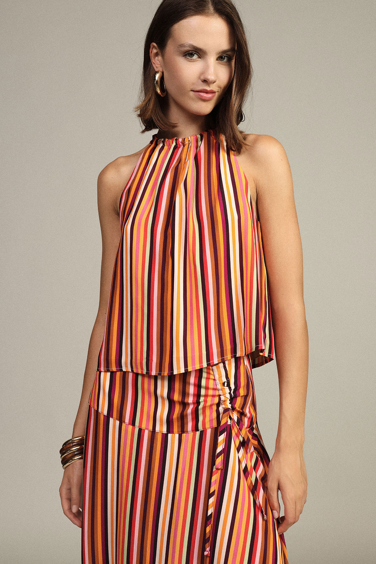 Woman wearing sleeveless halter dress with vertical multicolour stripes and side ruching, gold hoop earrings