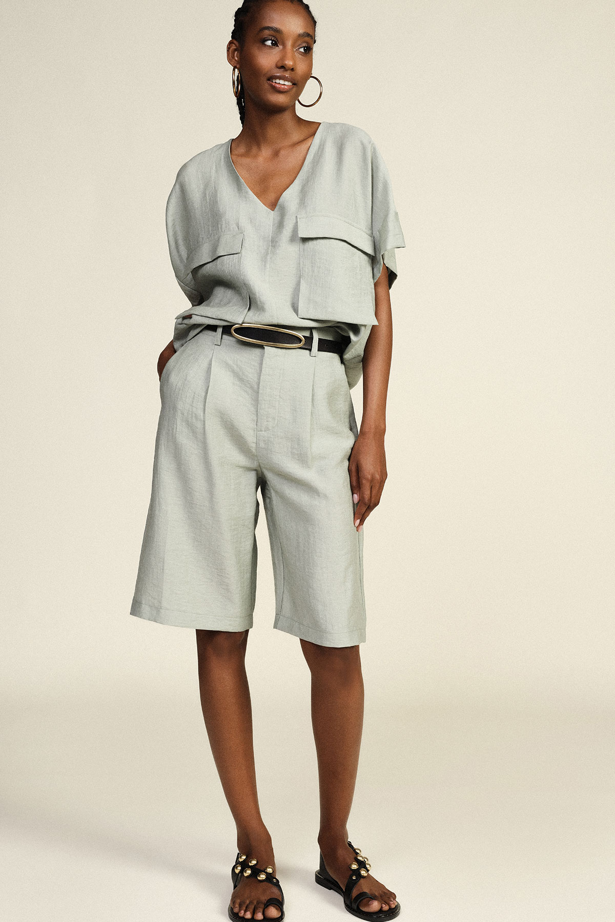 Model wearing sage-green linen V-neck top with oversized pockets and belted high-waist knee shorts, black stud sandals