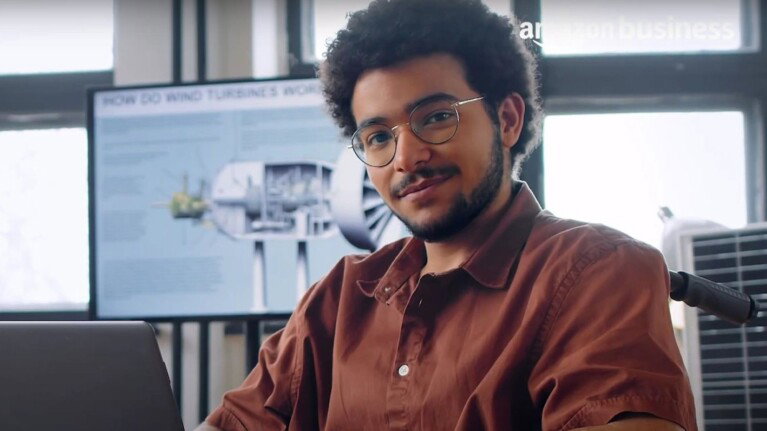 A man in glasses smiles beside his laptop in a bright office