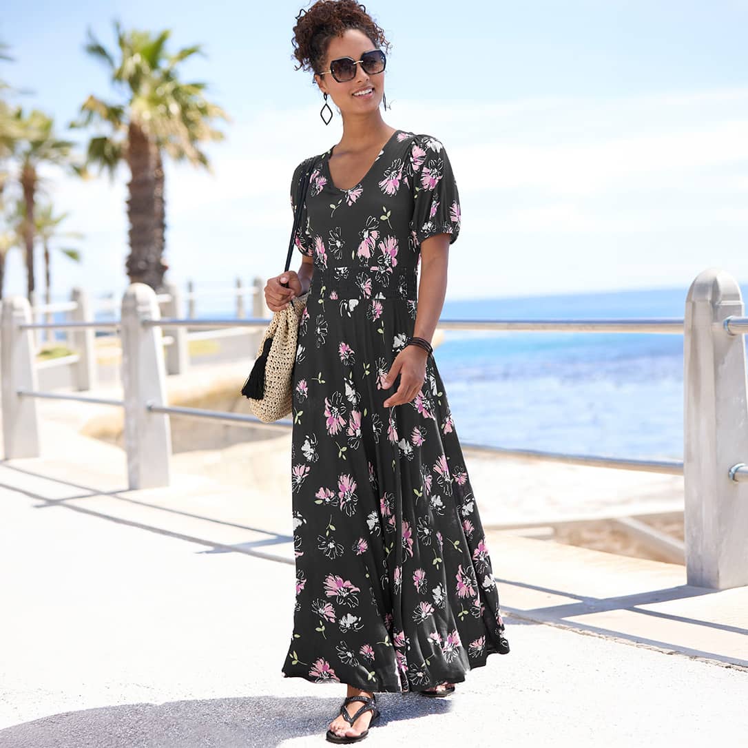 Ein Model mit lockigem Haar und Sonnenbrille trägt ein schwarzes, langes Kleid mit rosa Blumenmuster und geht am Meer entlang.
