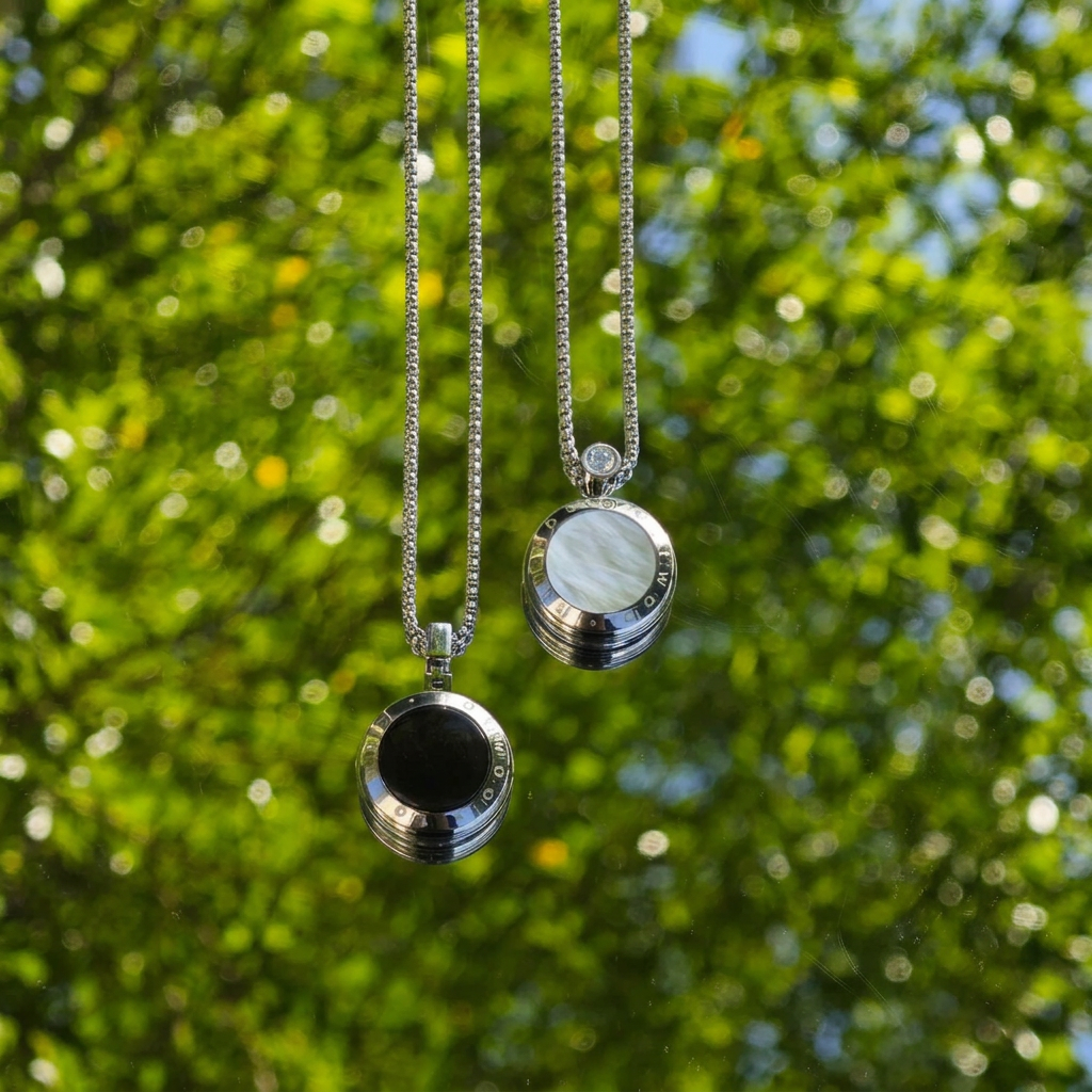 Smart Necklace Pair with silver crystal chains and matching pendants, one black stone and one pale stone, against soft green foliage