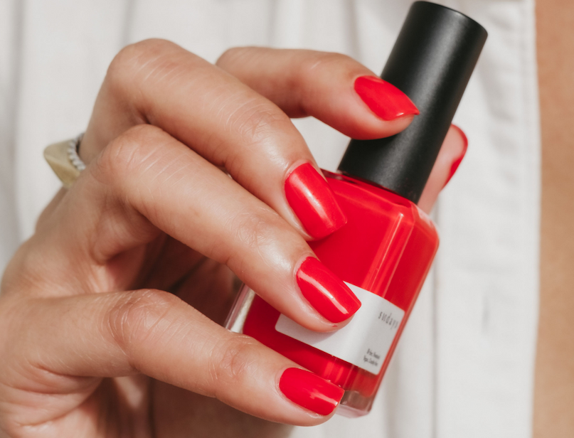 Hand with bright red nails holding a matching red nail polish bottle.
