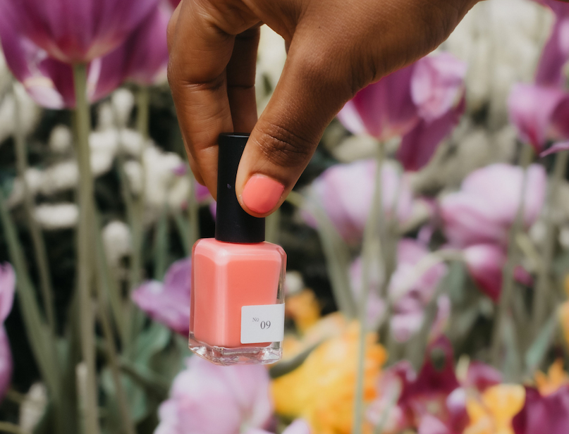 Hand holding a coral pink nail polish bottle in front of blooming flowers.