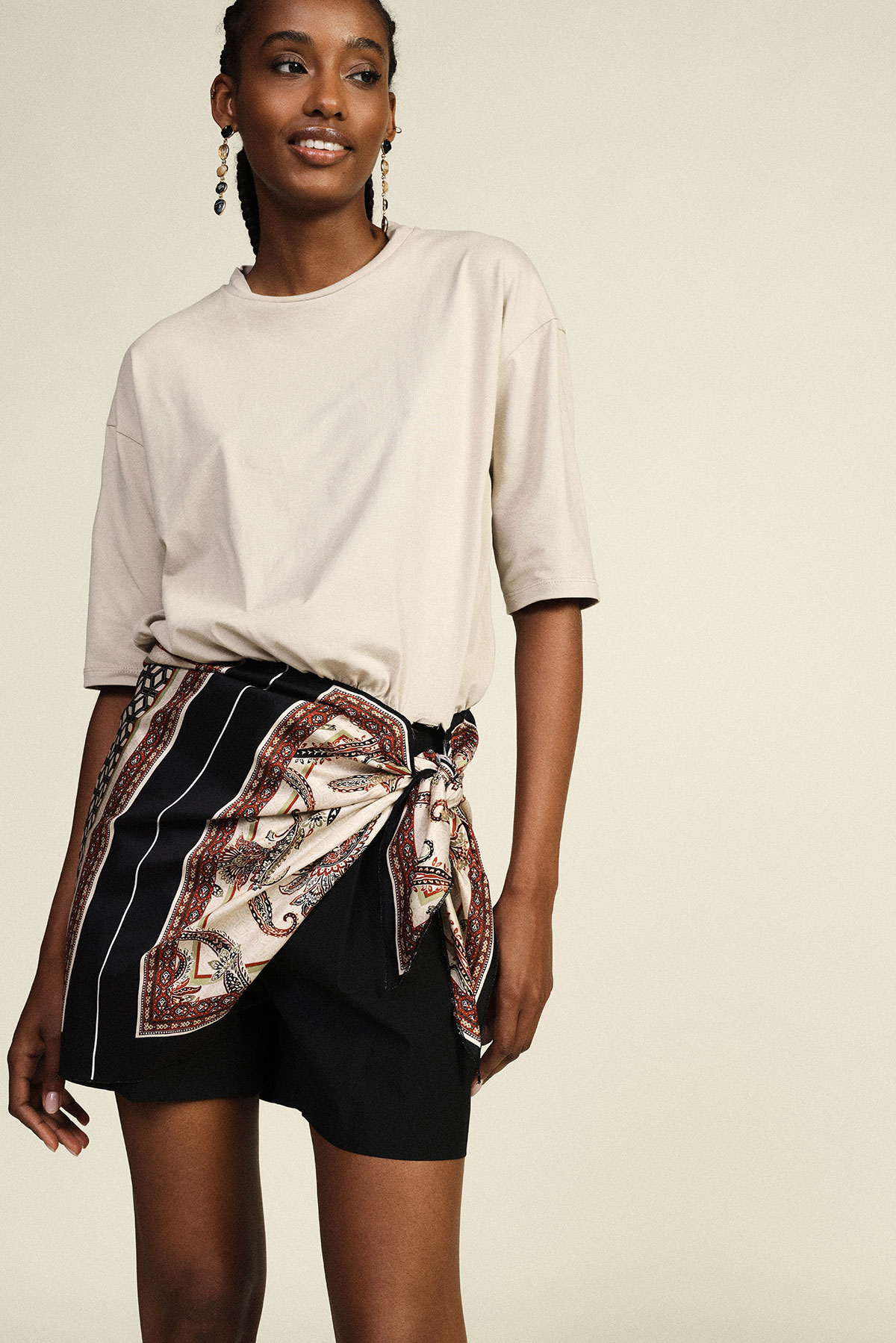Smiling woman modelling casual outfit: cream oversized T‑shirt, black shorts and patterned silk scarf tied at the waist, beaded drop earrings