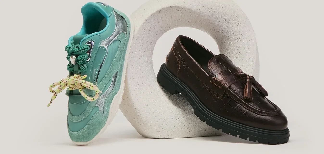 Baskets vertes avec lacets jaunes et mocassins en cuir marron foncé exposés sur et à côté d'une sculpture circulaire blanche sur un fond gris.