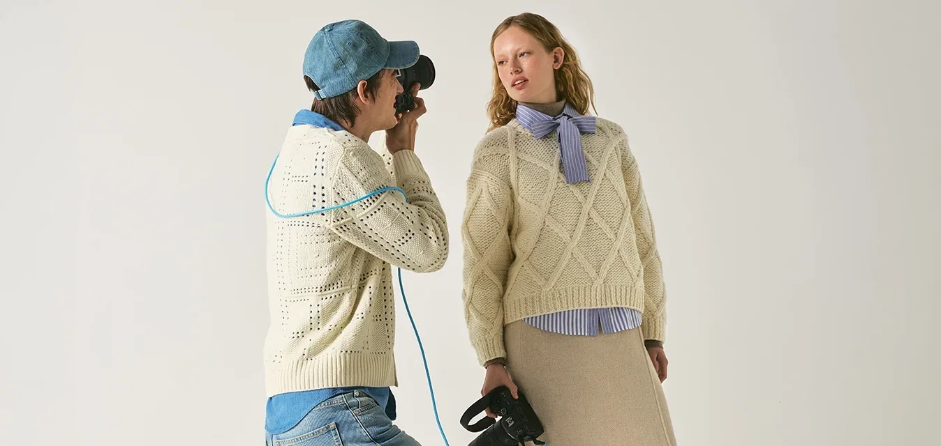 Photographe portant une casquette et un pull prend une photo d’une femme en pull en tricot et jupe beige tenant des bottes noires sur un fond uni.