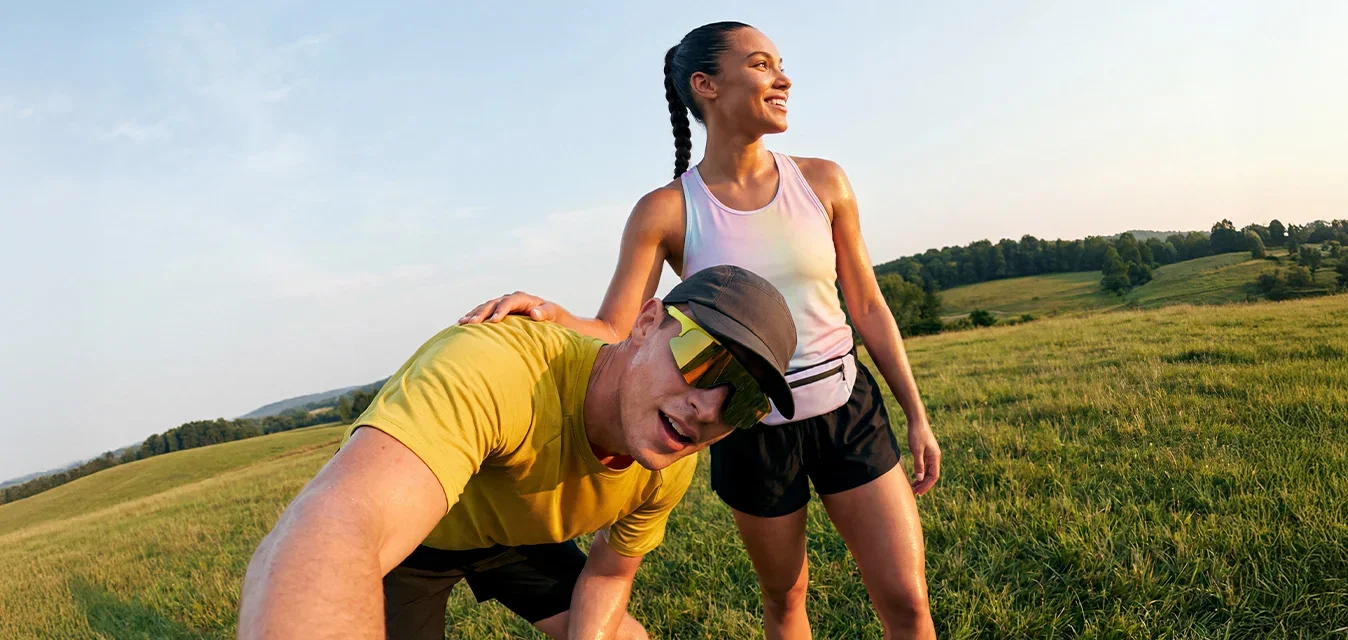 Deux coureurs se reposent dans un champ herbeux au coucher du soleil, l'un prenant un selfie tandis que l'autre sourit, tous deux vêtus de vêtements de sport.
