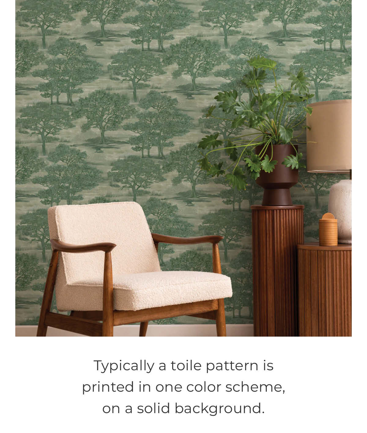 Image of Tempaper's Forest Toile wallpaper in green in a living space with a chair with a wood frame and tan cushions. Side tables and a lamp can be partially seen. Text below the image "Typically a toile pattern is printed in one color scheme, on a solid background. "
