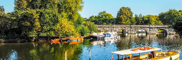 Boats moored by a tree-lined river with a stone bridge in the background under a clear blue sky.