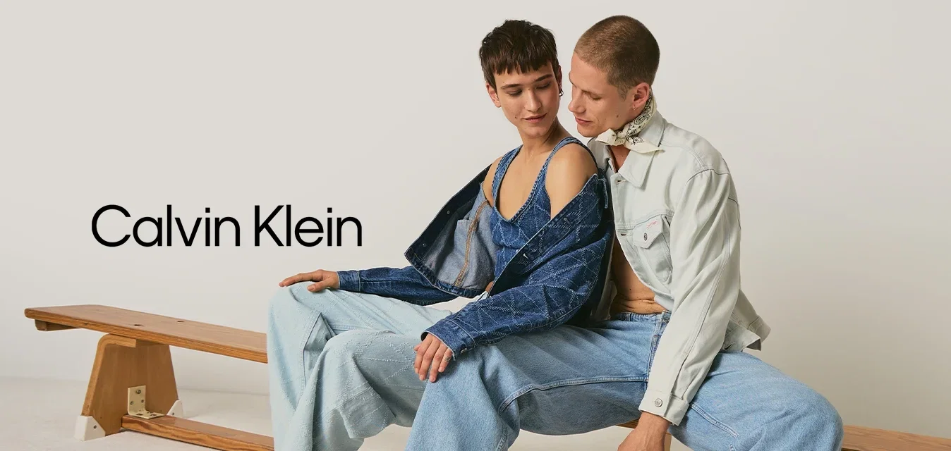 Twee jonge volwassenen, met blote voeten en in denim gekleed, zitten dicht naast elkaar op een houten bank tegen een effen achtergrond.