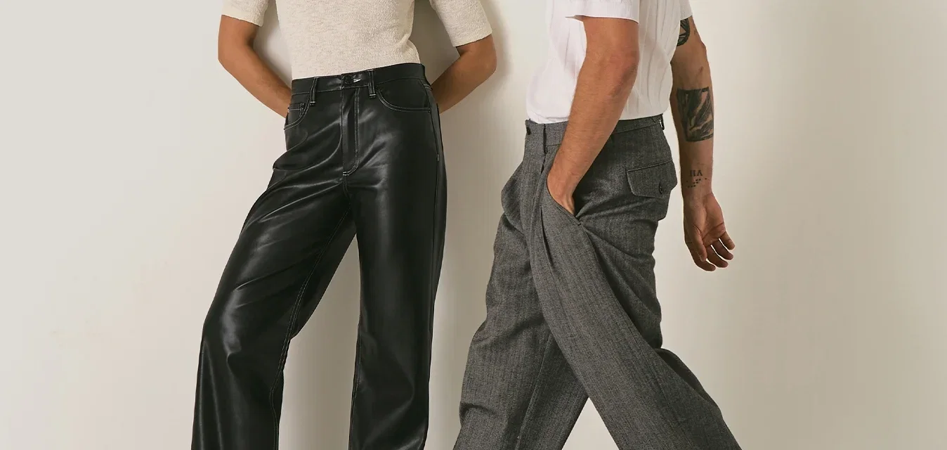 Twee stijlvolle volwassenen staan tegen een eenvoudige beige muur; vrouw in een wit shirt en zwarte broek, man in een wit shirt en grijze wijde broek aan het lopen.