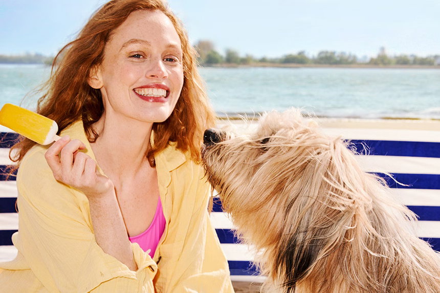 Eine Frau mit roten Haaren und gelber Bluse sitzt am Strand vor einem blauweiß gestreiften Windfang. Sie lächelt und hält ein gelbes Eis in der Hand. Vor ihr steht ein großer, langhaariger Hund, der sie neugierig anschaut.