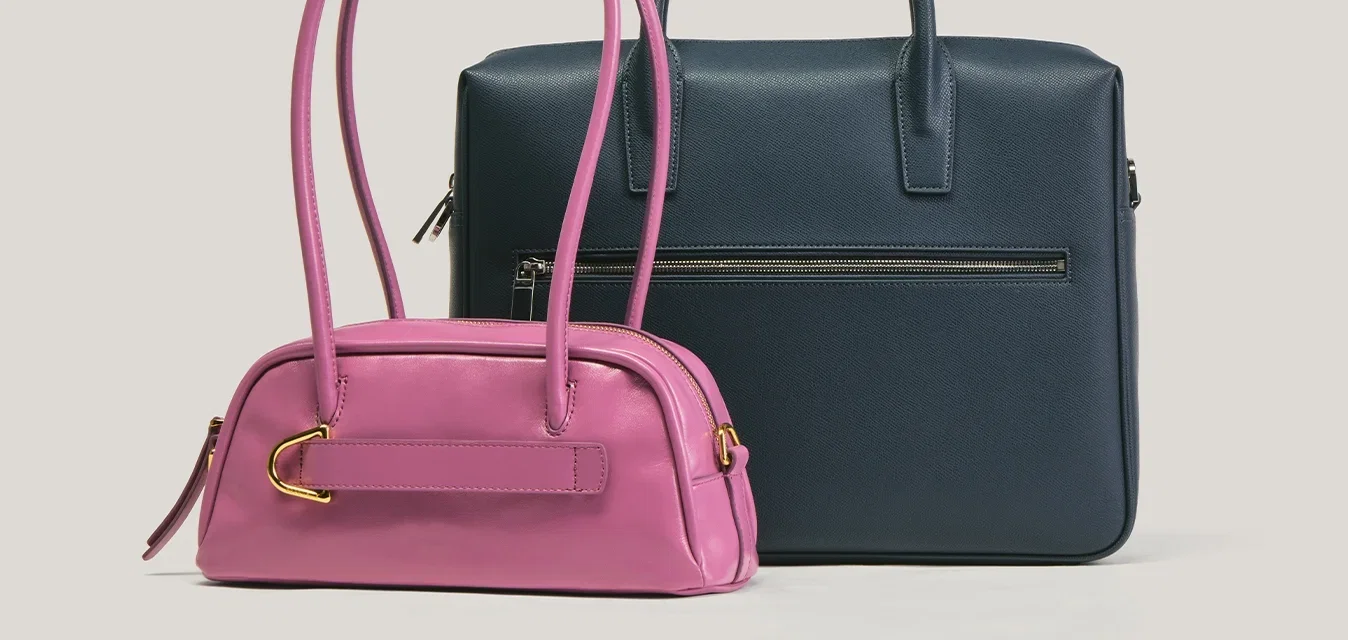 Pequeño bolso rosa con asas largas junto a un maletín más grande de estilo maletín azul oscuro con bolsillo frontal con cremallera sobre fondo liso.