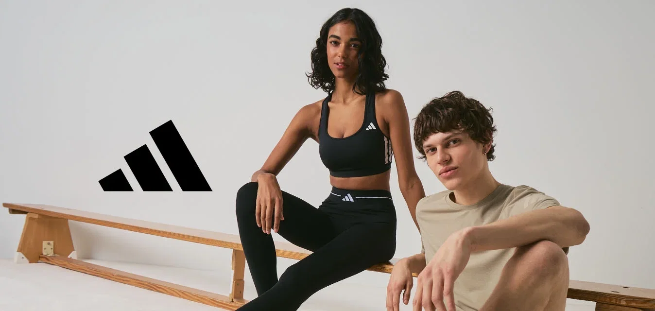 Dos adultos jóvenes con ropa deportiva están sentados cerca de bancos de madera en un espacio interior minimalista, ambos llevan zapatillas Adidas.