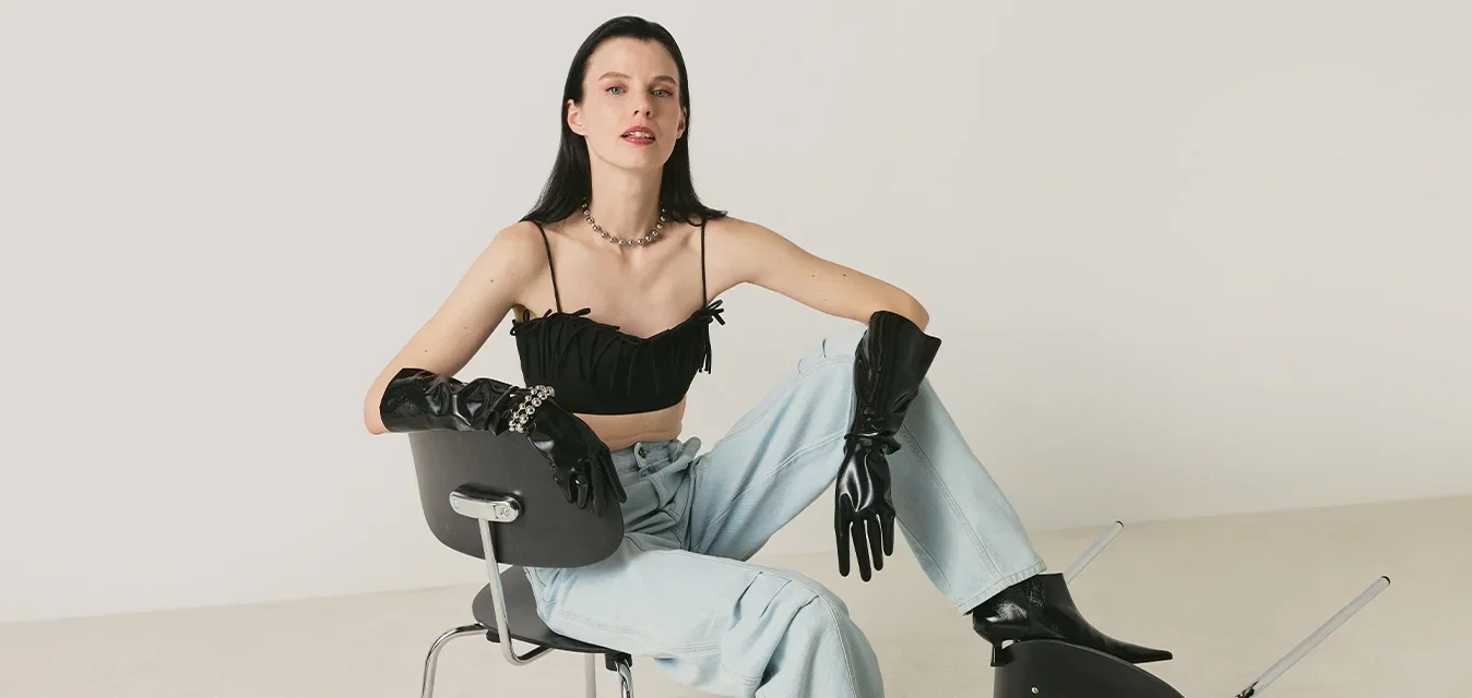 Joven mujer con top corto negro, guantes, botas y vaqueros azul claro, sentada en una silla con una pierna sobre una silla negra volcada.