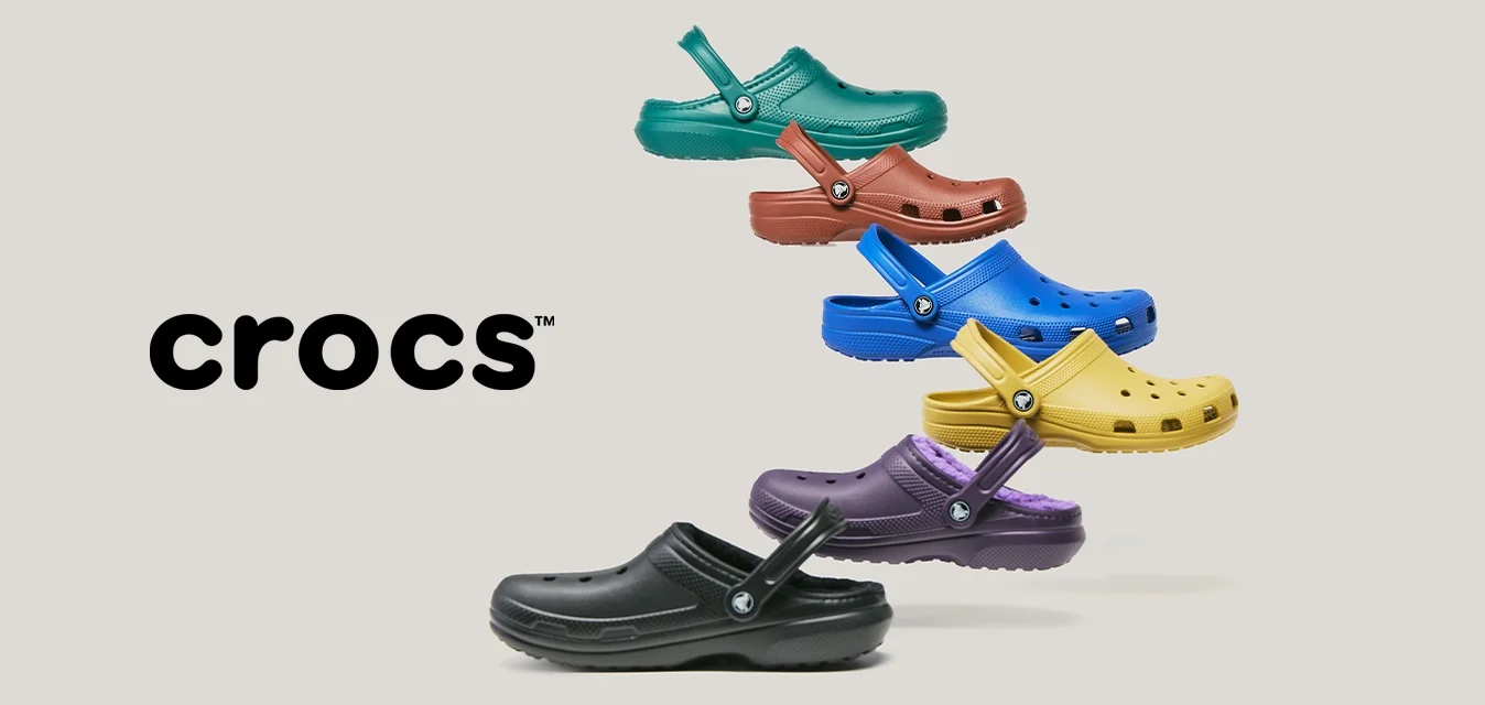 Seis coloridos zuecos Crocs en negro, púrpura, amarillo, azul, rojo y verde, dispuestos en una formación vertical escalonada sobre un fondo claro.