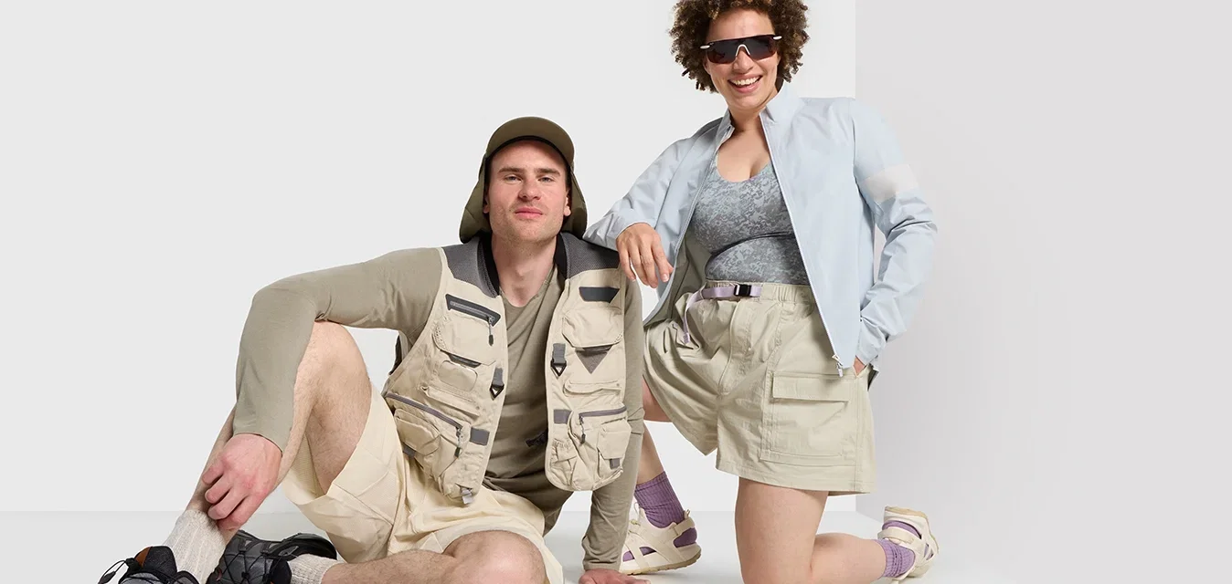 Dos personas con ropa de exterior: una con un chaleco de utilidad beige, camisa de manga larga gris y pantalones cortos; la otra con una chaqueta azul claro sobre una blusa con estampado.