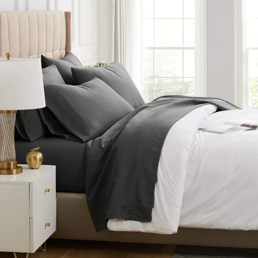 Modern bedroom with cream upholstered headboard, black and white layered bedding, nightstand with gold lamp and accessories, bright windows.