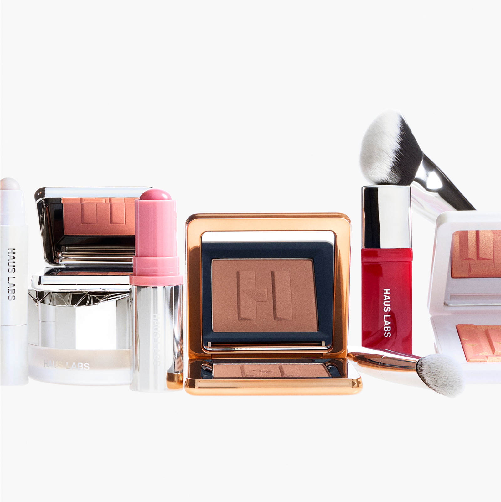 Assortment of Haus Labs complexion and color products, including blush and highlighter compacts, balm sticks, a red lip product, setting powder, and makeup brushes on a white background.