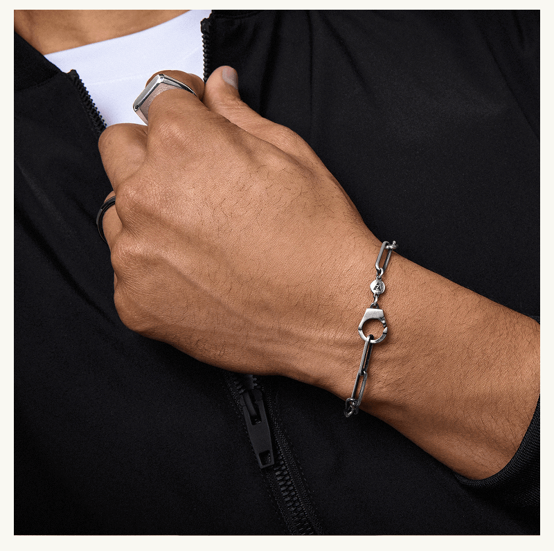 Male wearing silver chain bracelet with handcuff-shaped clasp & silver signet ring.