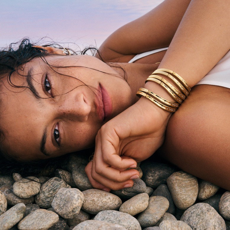 A model rests on pebbles wearing stacked 14k gold-plated bangles.