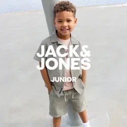 Jeune garçon souriant aux cheveux bouclés, portant une chemise gris clair et un short, un t-shirt rose, des chaussettes blanches et des baskets blanches, debout à l'extérieur près d'un poteau.